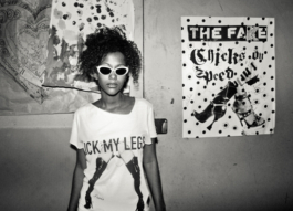 Photography. Black & white. Night clubber wears a punk t-shirt in front of poster cover wall. 